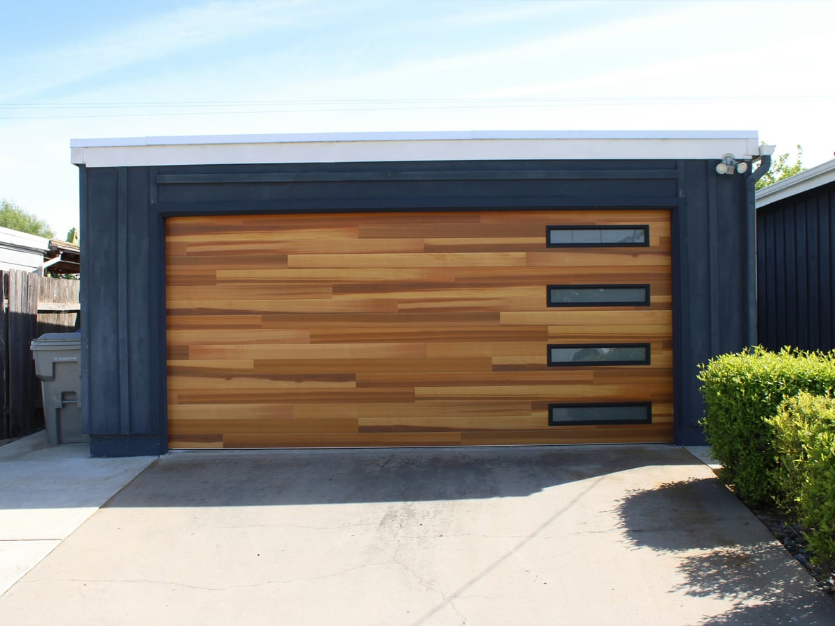 A modern wood-look garage door with four narrow windows on a dark-sided garage